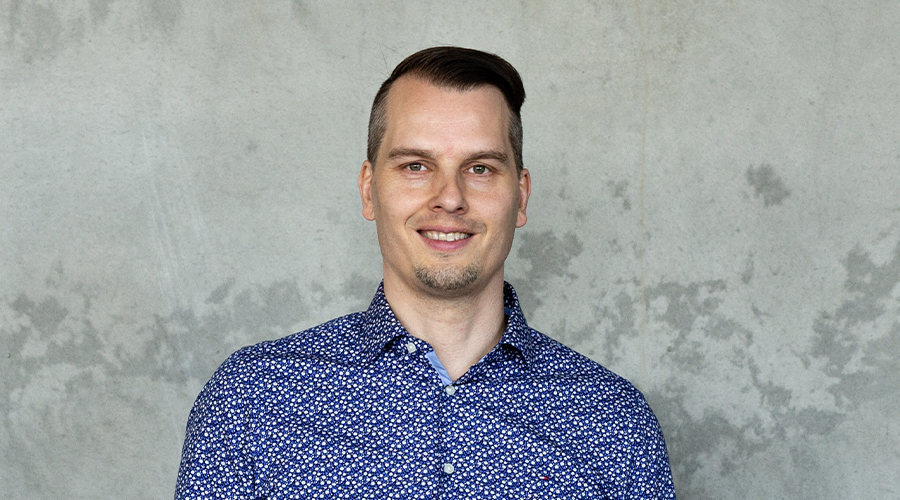 Kempeleen tekninen johtaja Tuomo Perälä sinisessä, kirjavassa kauluspaidassa kuvattuna harmaata betoniseinää vasten.