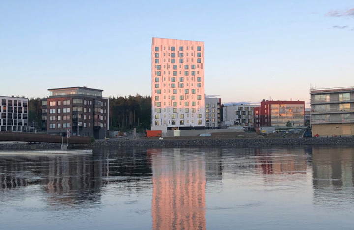 Kuvassa näkyy vaalea puukerrostalo järven rannalla muiisen, matalampien rakennusten välissä.