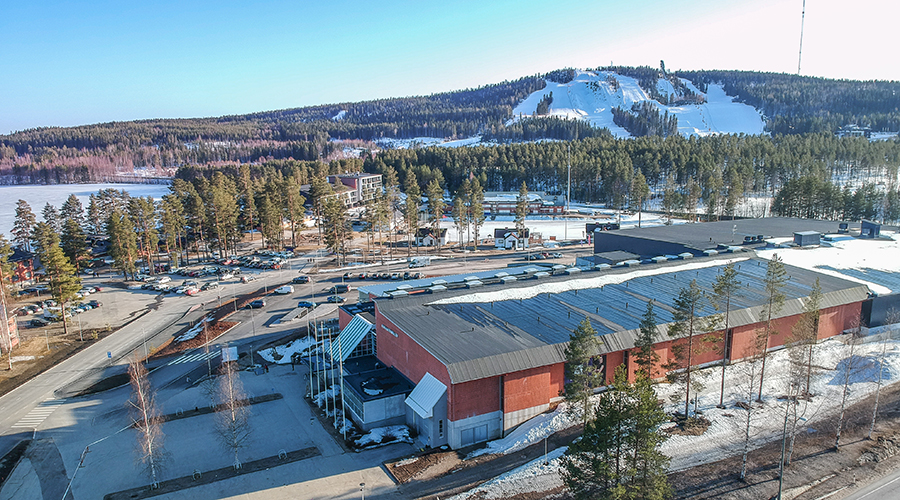 Vuokatti Areena ilmasta kuvattuna sinisen taivaan alla. Taustalla metsää ja Vuokatin laskettelurinteet.