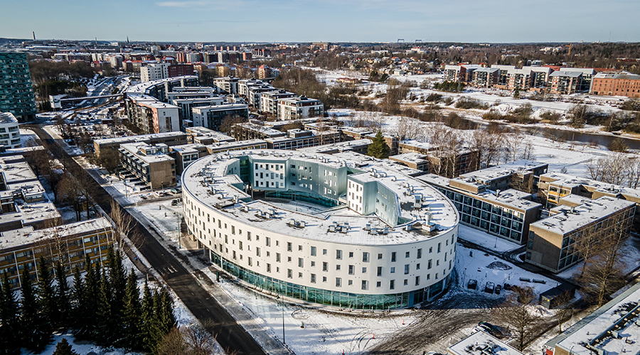 Ovaalinmuotoinen, valkoinen Tyyssija-rakennus ilmasta kuvattuna. Taustalla kaupunkia.